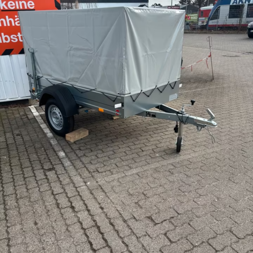 Silberner Stema PKW‑Planenanhänger FT 750 kg mit grauer Hochplane und Spriegel, einachsig auf Parkplatz abgestellt.